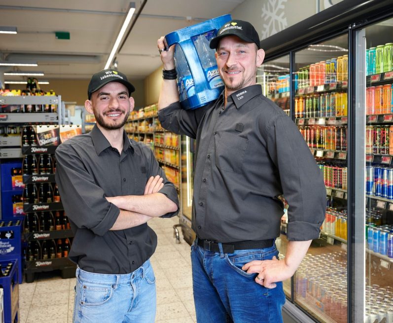 Abteilungsleitung Getränke Zwei Männer, einer hält eine Getränkekiste, Regale voller Getränke im Hintergrund.