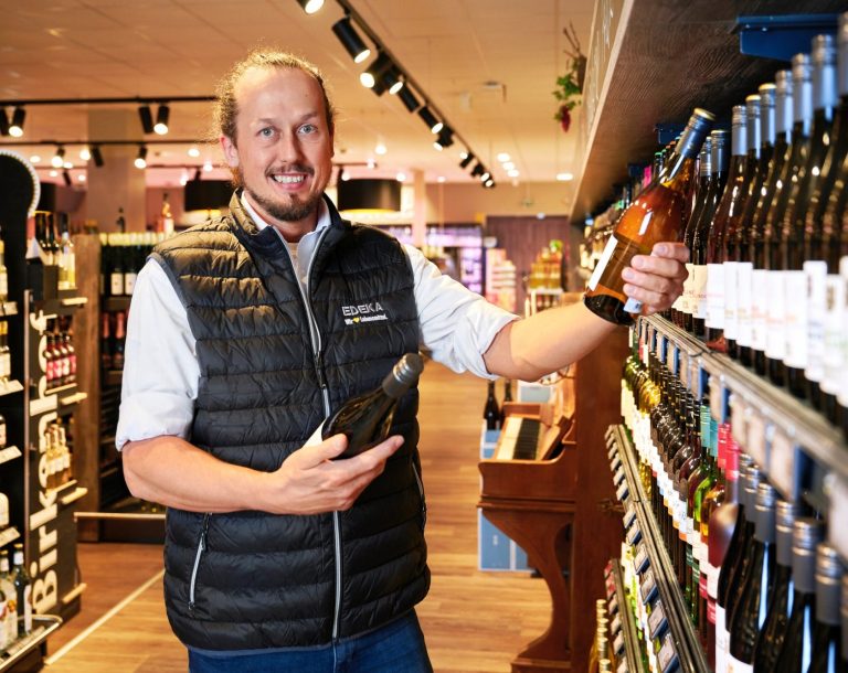 Marktleiter Mann in einem Weingeschäft hält eine Flasche Wein und zeigt auf Regale mit Weinflaschen.